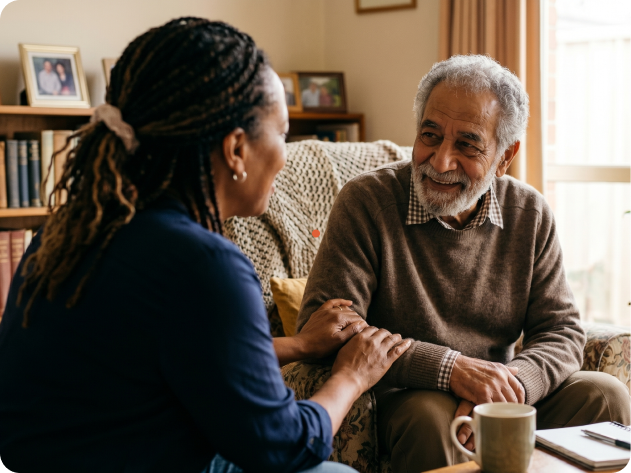 Woman and older man sit on a sofa, smiling and holding hands in a cozy living room, sharing a warm conversation.