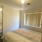 Small bedroom with a white bed and pastel-striped quilt, a white headboard, and a mirrored sliding-wardrobe on the left reflecting part of the bed. A window with vertical blinds is on the far wall.