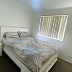 White wooden bed with tropical leaf-pattern duvet, two pillows, and a white nightstand in a small, bright bedroom.