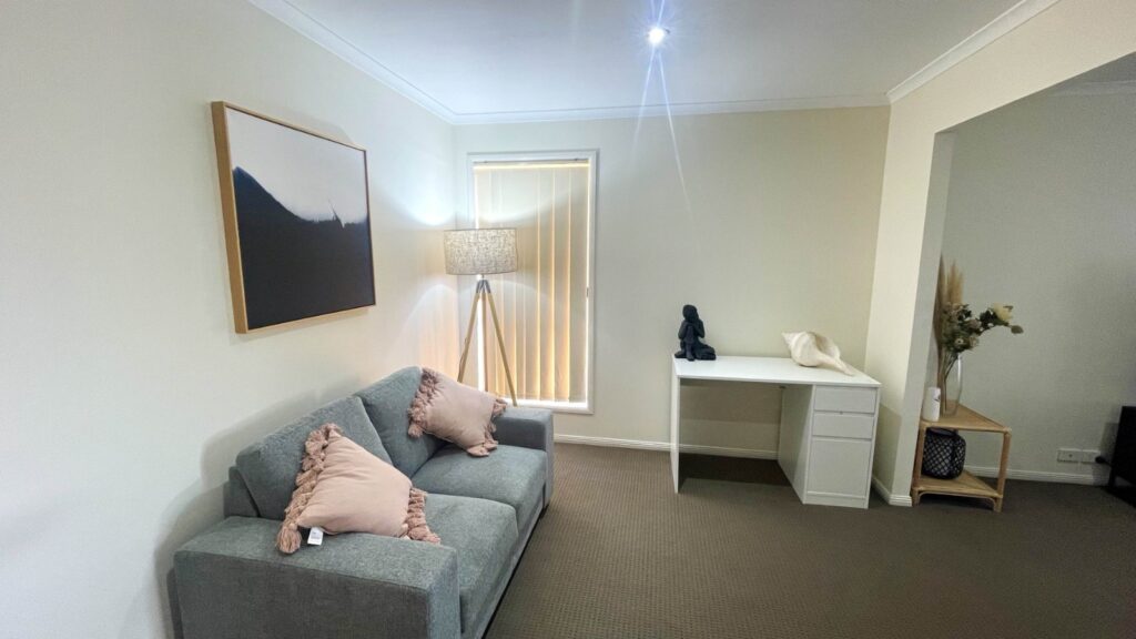 Living room with a gray sofa, blush-pink pillows, a framed mountain artwork on the wall, and a tripod floor lamp beside a window with vertical blinds.