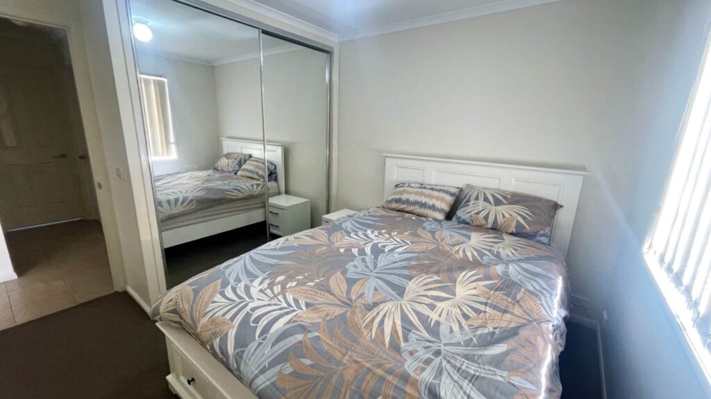 White wooden bed with a palm-patterned duvet in blue, beige, and gray, flanked by two small white nightstands in a bright bedroom