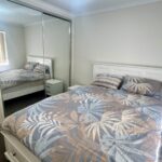 White wooden bed with a palm-patterned duvet in blue, beige, and gray, flanked by two small white nightstands in a bright bedroom