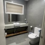 Gray-tiled bathroom with a wall-mounted white sink on a long counter, a large mirror, and a white toilet nearby.