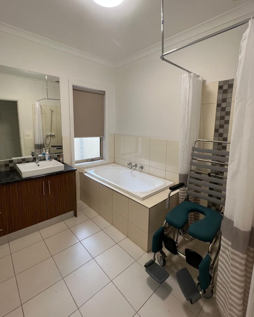 Bathroom with a white bathtub, sink vanity, and large mirror; a wheeled bath chair with teal cushions is beside a striped shower curtain.