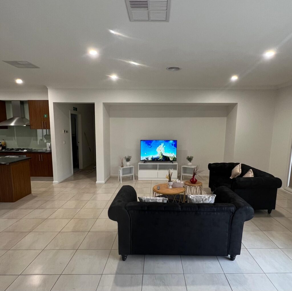 Open-plan living room with black sectional sofas facing a TV on a white stand; kitchen visible to the left.