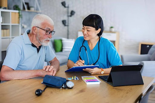 Nurse in blue scrubs helps an elderly man at a table, reviewing a clipboard and medical supplies.