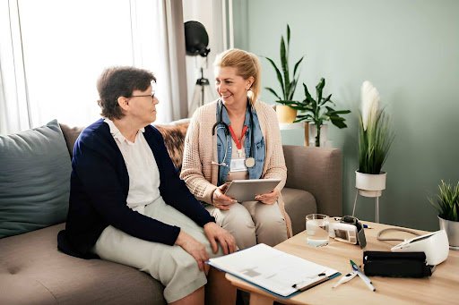 Healthcare professional with a stethoscope sits with an elderly woman on a living room couch, discussing with a tablet nearby.