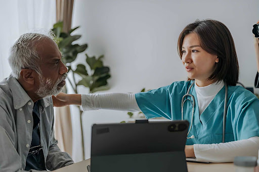 Female healthcare professional in teal scrubs comforting an elderly man during a medical consultation in a bright clinic.