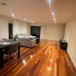 Open-plan living and dining area with a dark wood dining table and six gray chairs, gray sofas, and warm recessed lighting on wood floors at night in a spacious room. l