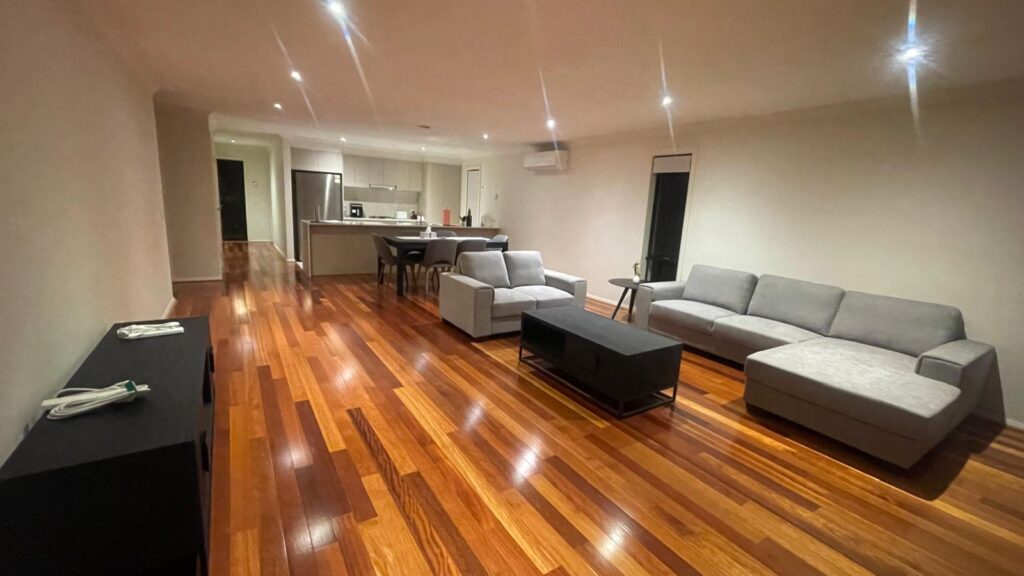 Open-plan living area with a kitchen in the background: hardwood floors, gray couches, a black coffee table, and a dining table nearby.