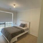 Small white bedroom with a white bed frame, gray bedding, and a window with a rolled-down blind across from the bed.