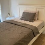 Neutral-toned bedroom with a white paneled bed frame and gray bedding, including a gray pillow and matching blanket. A white nightstand holds a small vase and a cup.