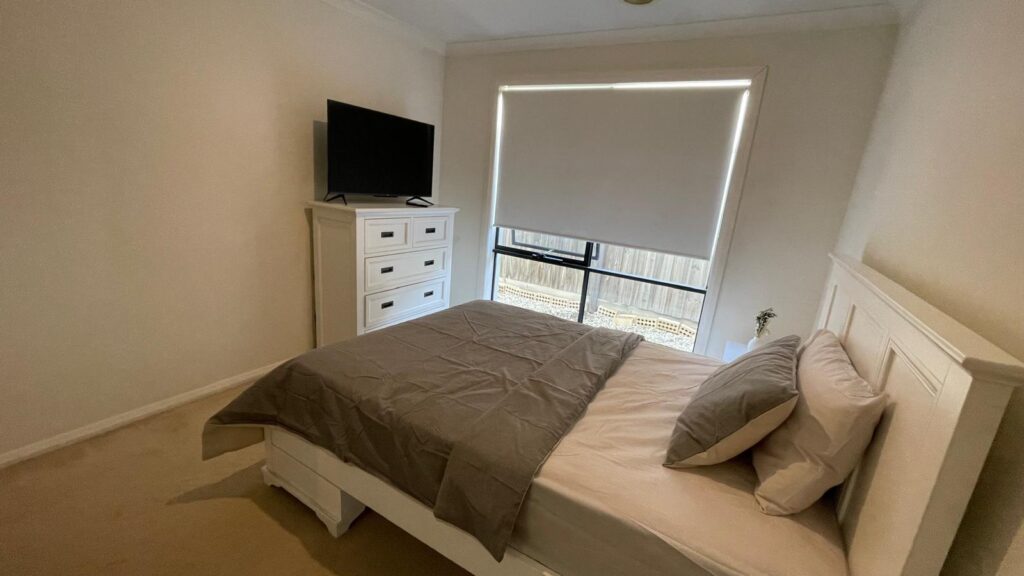 Modern white bedroom with a double bed, gray bedding, and a white headboard; a dresser with a TV on top sits nearby.