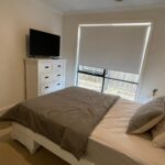 Modern white bedroom with a double bed, gray bedding, and a white headboard; a dresser with a TV on top sits nearby.