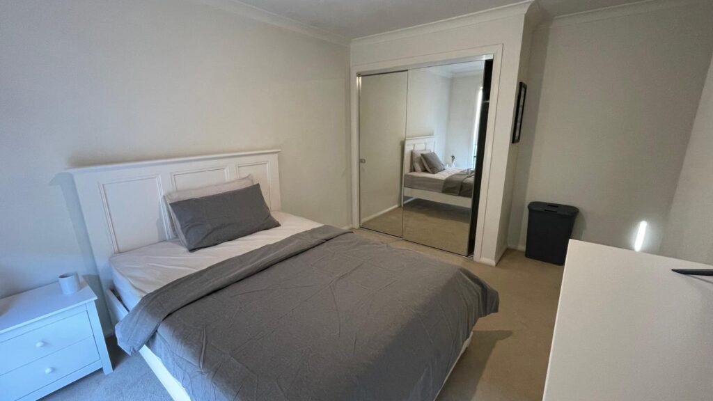Modern bedroom with a white paneled bed in gray sheets, a matching gray pillow and blanket, white nightstand, and a mirrored sliding-door wardrobe reflecting the bed. A white dresser and a dark laundry bin sit to the side.