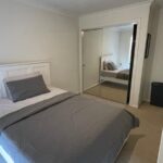 Modern bedroom with a white paneled bed in gray sheets, a matching gray pillow and blanket, white nightstand, and a mirrored sliding-door wardrobe reflecting the bed. A white dresser and a dark laundry bin sit to the side.